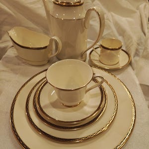Puede incluir: Un juego de té color crema con ribetes dorados, que incluye una tetera, una crema, una taza y un platillo, y platos. El juego tiene un diseño clásico con un borde negro y dorado. El juego de té está dispuesto sobre una superficie blanca.