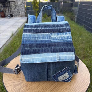 May include: A backpack constructed from various shades of blue denim fabric featuring a patchwork design on the flap. The bag includes a top handle, adjustable gray shoulder straps, and a small rectangular label on the front that reads New Look Kim Tori.