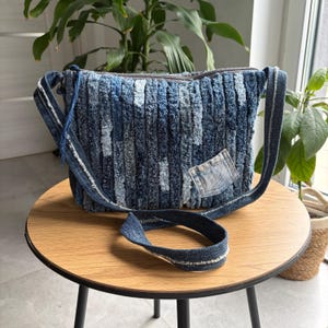 May include: A blue denim shoulder bag with a textured, vertical stripe design. The bag has a zip closure, a denim strap, and a small denim patch. The bag is on a round wooden table.