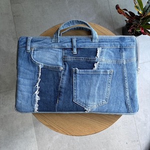 May include: A rectangular denim laptop bag with a handle, made from repurposed jeans. The bag features a patchwork design with various shades of blue denim, including pockets and distressed details. The bag is sitting on a wooden table.