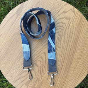 May include: A long, adjustable strap made from various shades of blue denim fabric patches sewn together in a patchwork design. The strap features grey webbing at both ends, each finished with a silver-toned metal swivel lobster clasp hook.