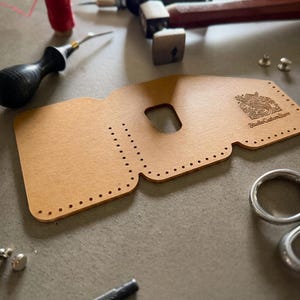 Può includere: Un modello per portafoglio in pelle color cuoio con fori di cucitura pre-perforati, un ritaglio centrale e il logo stampato StudioCustomStore. Il pezzo è circondato da strumenti per la lavorazione della pelle tra cui martello, lesina, filo rosso e rivetti.