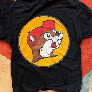 May include: A black short-sleeved t-shirt featuring a large circular graphic on the chest. The graphic displays a brown beaver character wearing a red baseball cap, set against a solid yellow background with a distressed, weathered texture.