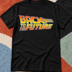 May include: A black short-sleeved t-shirt featuring the Back to the Future logo printed on the chest. The graphic text displays a distressed, weathered texture with a color gradient transitioning from yellow to orange, outlined in light blue.