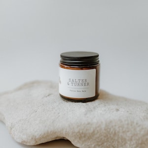May include: A small, brown glass jar with a black lid, labelled "SALTER & TURNER Yellow Skin Balm." The jar rests on a light-coloured, textured stone. The background is a plain, off-white colour.