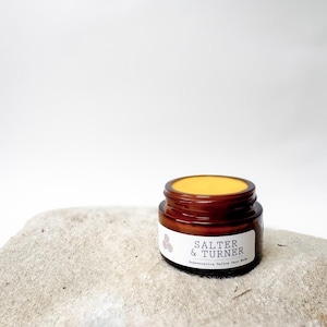 Op de afbeelding: Een klein, bruin glazen potje gezichtsbalsem met een gele bovenkant. Het potje heeft een wit etiket met de tekst "SALTER & TURNER" en "Regenerative Tallow Face Balm". Het potje staat op een grijze steen.