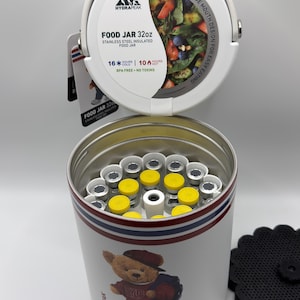 Puede incluir: Un termo blanco aislado para alimentos con una capacidad de 946 ml, con un oso de peluche y rayas rojas, blancas y azules. La tapa está abierta, revelando el interior y el texto "FOOD JAR 32oz". Incluye una base negra en forma de flor.