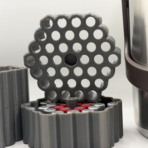 May include: A gray 3D-printed cartridge holder with a honeycomb-like top, holding several silver and red cartridges. A stainless steel container with a brown handle is visible in the background. The holder is designed to organize and store items.