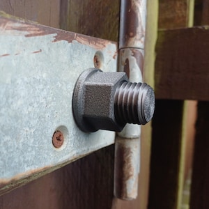 May include: A dark grey, textured, hexagonal nut with a threaded bolt end protruding from its centre. The nut is mounted onto a flat, rectangular, galvanised metal plate that is secured with a visible screw against a brown wooden surface.