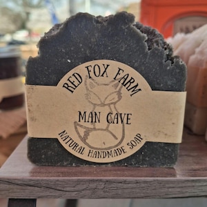 May include: A dark gray, textured bar of handmade soap with a paper label. The label features a fox illustration and the words "Red Fox Farm," "Man Cave," and "Natural Handmade Soap."