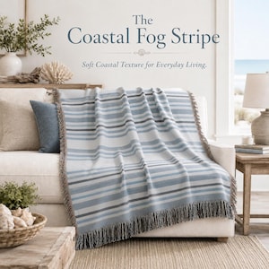 May include: A woven throw blanket featuring horizontal stripes in shades of light blue, grey, and white with fringed edges. The text "The Coastal Fog Stripe" and "Soft Coastal Texture for Everyday Living" is printed above the blanket.