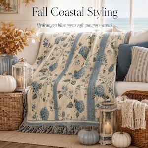 May include: A woven throw blanket featuring a cream background with vertical blue stripes and a floral hydrangea pattern. The blanket has a fringed edge and is displayed draped over a sofa. Text on the image reads Fall Coastal Styling and Hydrangea blue meets soft autumn warmth.