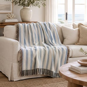 May include: A blue and white striped throw blanket with fringed edges is draped over a white sofa. The blanket's vertical stripes alternate between light blue and white. The fringed edges have a mix of brown and black threads. The sofa is in a living room setting.