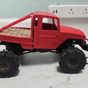 May include: A red toy truck with large black tires. The truck has a cab and a flatbed with a wooden-like texture. The truck has a roll bar and black suspension. The toy truck is a miniature replica.