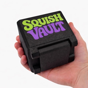 May include: A black box with the text "SQUISH VAULT" in green and purple lettering is held in a hand. The box has a textured surface and a hinged lid. The background is white.