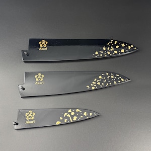 以下が含まれることがあります： 金色のアクセントが付いた黒いナイフシース3つ。各シースには金のフローラルデザインと「Akari」の文字があります。金色の葉のようなディテールが先端を飾っています。シースは濃い灰色の表面に配置されています。