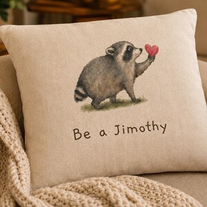 May include: A square beige throw pillow featuring a printed illustration of a raccoon holding a small red heart. Below the raccoon, the text "Be a Jimothy" is printed in a simple brown font. The pillow has a textured, woven fabric appearance.