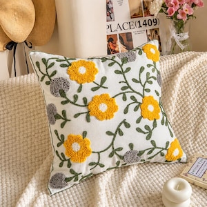 May include: A square throw pillow with a white fabric base featuring a tufted floral pattern. The design includes bright yellow flowers with white centers, grey circular tufted accents, and dark green embroidered vines and leaves.