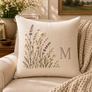 May include: A square off-white throw pillow featuring an embroidered design of wildflowers, including white daisies and purple lavender sprigs, on the left side. The letter M is embroidered in a simple serif font on the right side of the pillow.