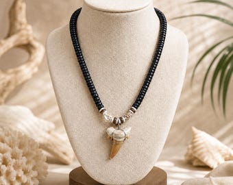 May include: A black beaded necklace with a large, light-colored shark tooth pendant. The necklace is displayed on a beige mannequin bust. The shark tooth is a light tan color with a cream-colored base. The necklace also features small shell accents.