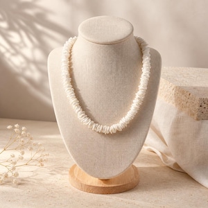 May include: A white shell necklace displayed on a beige jewelry bust with a wooden base. The necklace is made of small, irregular shell pieces strung together. The background is a neutral beige with soft shadows.