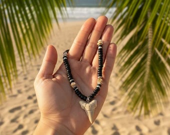 May include: A black beaded necklace with a shark tooth pendant. The necklace features black and white beads, with shell accents. The shark tooth is a light beige color. The background shows a beach scene with palm leaves.