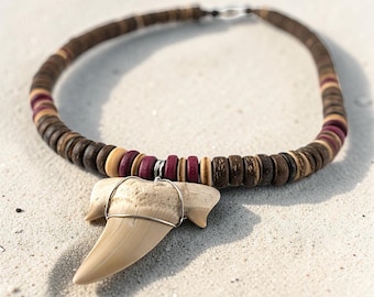 May include: A necklace featuring a large, off-white shark tooth pendant, secured with silver wire, hangs from a strand of brown, tan, and burgundy wooden beads. The necklace rests on a sandy surface, suggesting a beach theme.