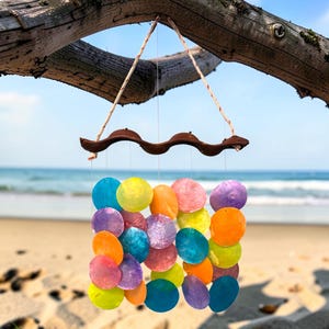 May include: A vibrant seashell wind chime suspended from a tree branch. The chime has a wooden top with a curved design and is adorned with a collection of brightly coloured shells in blue, yellow, orange, pink, and purple.