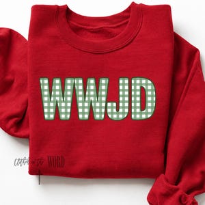 Può includere: Una felpa rossa a maniche lunghe con girocollo caratterizzata dalle lettere WWJD in un motivo a quadretti vichy verde e bianco. Le lettere hanno un contorno verde scuro. Il testo Clothed in the WORD è stampato in piccole lettere nere in basso.