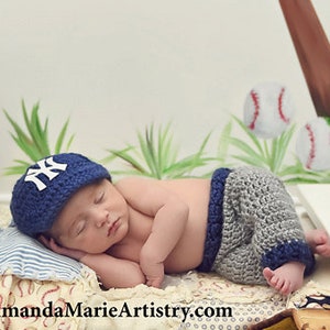 Pu&ograve; includere: Un berretto da baseball all'uncinetto blu navy con logo NY bianco e pantaloncini all'uncinetto abbinati grigi e blu navy. Il set &egrave; esposto su un piccolo letto in legno bianco con una trapunta a tema baseball, un guanto in pelle e una palla.