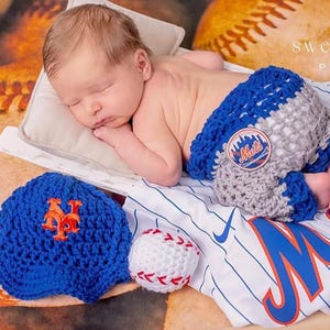 Pu&ograve; includere: Un neonato indossa copripannolino all'uncinetto blu e grigio con una toppa del logo Mets. Il set include un berretto da baseball all'uncinetto blu con logo NY arancione, una pallina da baseball all'uncinetto bianca e una maglia bianca a righe blu.