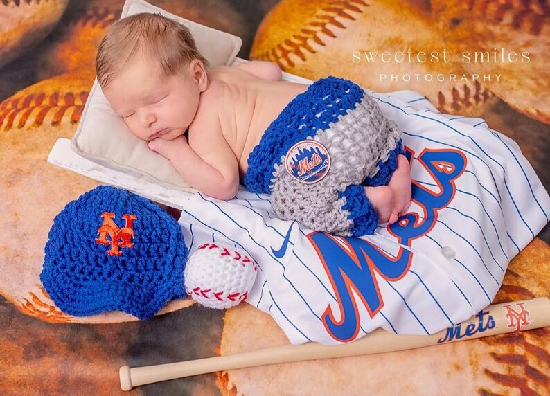 Pu&ograve; includere: Un neonato indossa copripannolino all'uncinetto blu e grigio con una toppa del logo Mets. Il set include un berretto da baseball all'uncinetto blu con logo NY arancione, una pallina da baseball all'uncinetto bianca e una maglia bianca a righe blu.