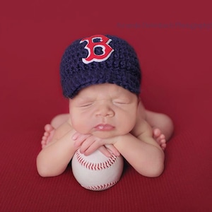 Può includere: Un bambino indossa un berretto da baseball blu con una "B" rossa e dorme con la testa appoggiata su una palla da baseball bianca. Il bambino è sdraiato su una superficie rossa.