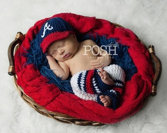 Può includere: Un berretto da baseball all'uncinetto blu navy con una lettera A bianca sul davanti e pantaloni all'uncinetto abbinati con strisce orizzontali blu navy, bianche e rosse. Il set è esposto all'interno di un cesto intrecciato foderato con tessuto rosso e blu.