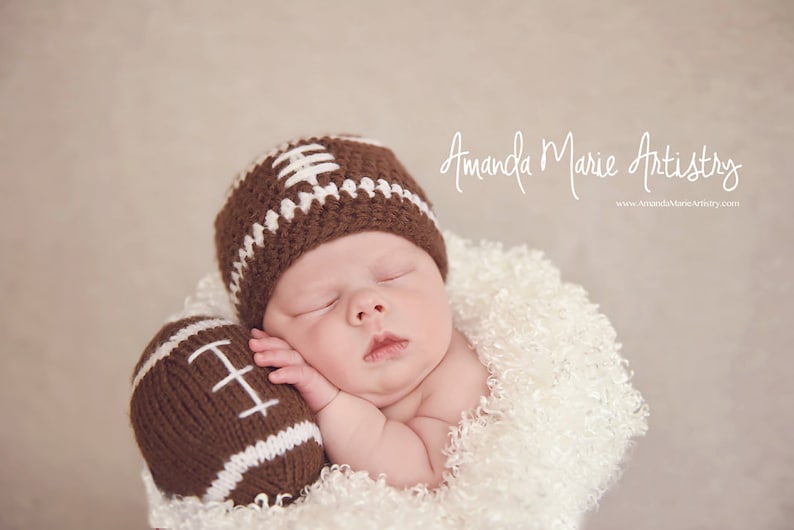 May include: A sleeping baby wearing a brown and white knitted football hat and holding a brown and white knitted football. The baby is lying on a white fluffy blanket.