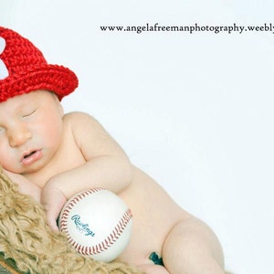 May include: A sleeping baby wearing a red crocheted baseball cap with a white "P" on it, holding a white baseball with red stitching and the word "Rawlings" printed on it.