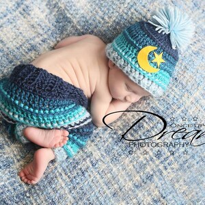 May include: A sleeping baby wearing a blue and white crocheted hat with a yellow crescent moon and star design. The baby is also wearing a blue and white crocheted pants.