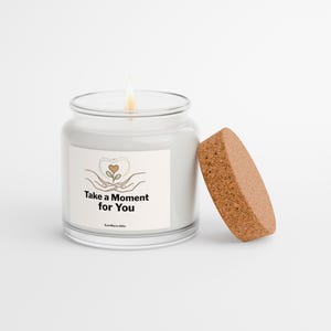 May include: A clear glass candle jar with a lit wick and a separate cork lid. The label features a line drawing of hands holding a small plant with a heart-shaped leaf and the text Take a Moment for You and EverWarm Gifts.