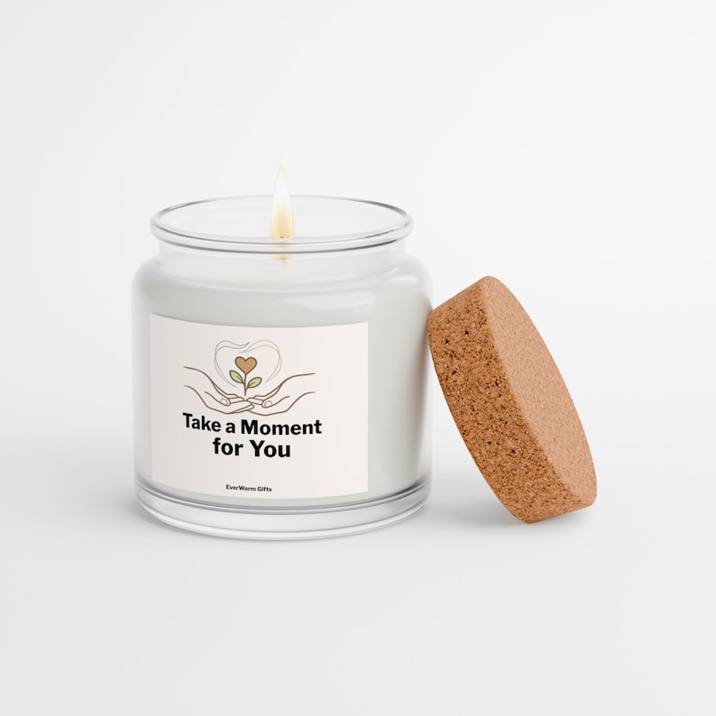May include: A clear glass candle jar with a lit wick and a separate cork lid. The label features a line drawing of hands holding a small plant with a heart-shaped leaf and the text Take a Moment for You and EverWarm Gifts.