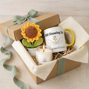 May include: A gift box containing a crocheted sunflower in a white pot, a white ceramic mug with a yellow handle featuring the text Still Showing Up and an illustration of hands holding a flower, and a scented candle with a white label.