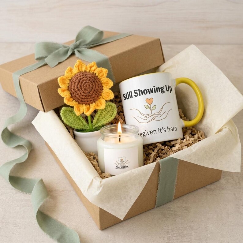 May include: A gift box containing a crocheted sunflower in a white pot, a white ceramic mug with a yellow handle featuring the text Still Showing Up and an illustration of hands holding a flower, and a scented candle with a white label.