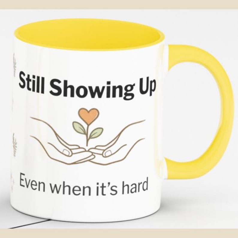 May include: A white ceramic mug with a bright yellow interior and handle. The front features the black text Still Showing Up above a line drawing of two hands cradling a small plant with a heart-shaped leaf, followed by the text Even when it's hard.
