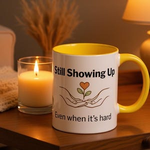 May include: A white ceramic mug with a bright yellow interior and handle. The front features the black text Still Showing Up and Even when it's hard, with a graphic of two hands cupping a small plant with an orange heart-shaped leaf in the centre.