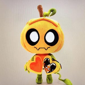 Pode incluir: Um personagem de ab&oacute;bora de pel&uacute;cia com olhos brancos grandes, contornos pretos e uma boca costurada. Ele segura um objeto em forma de cora&ccedil;&atilde;o com um cora&ccedil;&atilde;o laranja em um lado. A pel&uacute;cia tem pernas verdes e um caule.