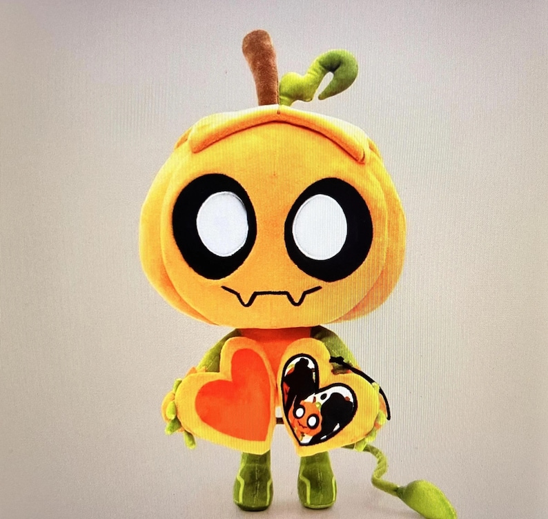 Pode incluir: Um personagem de ab&oacute;bora de pel&uacute;cia com olhos brancos grandes, contornos pretos e uma boca costurada. Ele segura um objeto em forma de cora&ccedil;&atilde;o com um cora&ccedil;&atilde;o laranja em um lado. A pel&uacute;cia tem pernas verdes e um caule.