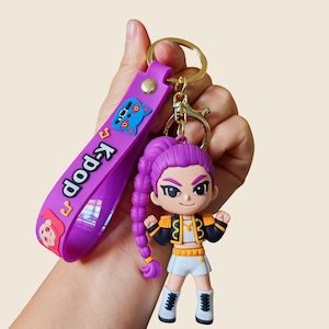 May include: A purple K-Pop keychain featuring a cartoon character with purple hair, wearing a military-style jacket and white shorts. The keychain has a purple strap with the text "K-Pop" and a cartoon cat design.