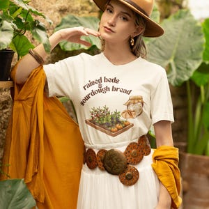 May include: A woman wearing a tan fedora, a white t-shirt with the text "raised beds & sourdough bread", and a wide brown belt with circular embellishments. She is also wearing a white dress and a mustard-coloured shawl.
