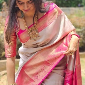 May include: A beige saree featuring a vibrant hot pink border with intricate gold zari woven patterns. The matching blouse has short sleeves with gold embroidery. The ensemble is paired with a large gold pendant necklace and stacked pink bangles.