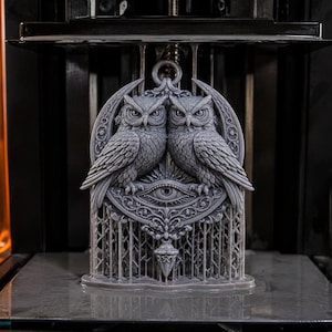 Pode incluir: Uma escultura de resina cinza impressa em 3D apresentando duas corujas detalhadas empoleiradas lado a lado sobre um motivo de olho central, emolduradas por uma lua crescente e padrões de filigrana, apoiadas por uma estrutura de grade vertical.