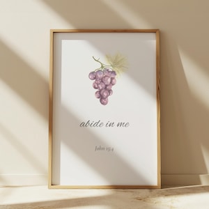 Puede incluir: Una impresión enmarcada con una ilustración de acuarela de uvas moradas y hojas verdes. El texto "abide in me" y "John 15:4" está escrito en una delicada escritura debajo de las uvas. El marco es de madera clara.